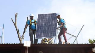 Two young men wearing hard hats and fall protection harnesses get ready to place a solar panel on the roof of a building