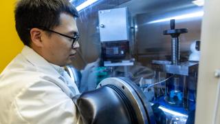 Wearing a white lab coat, postdoc researcher Yaohong Xiao reaches into a isolated chamber to work on battery research