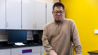 Associate Professor of Industrial and Manufacturing Systems Engineering Jian Hu stands for a portrait in his lab, with his hands on a benchtop and storage cabinets in the background.