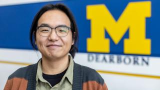 Health and Human Services Assistant Professor Vitalis Im poses for a head-and-shoulders portrait in front of a UM-Dearborn logo painted on wall. 