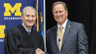 Two men, one wearing a judges robe, shake hands while standing for a portrait in front of a UM-Dearborn-themed background.